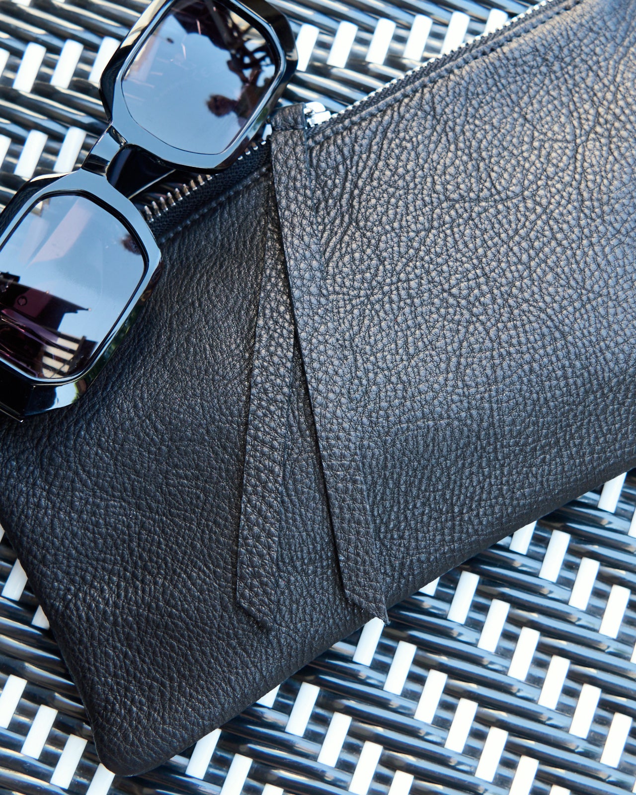 A black leather zip pouch, styled with black sunglasses on a black and white woven patio chair.